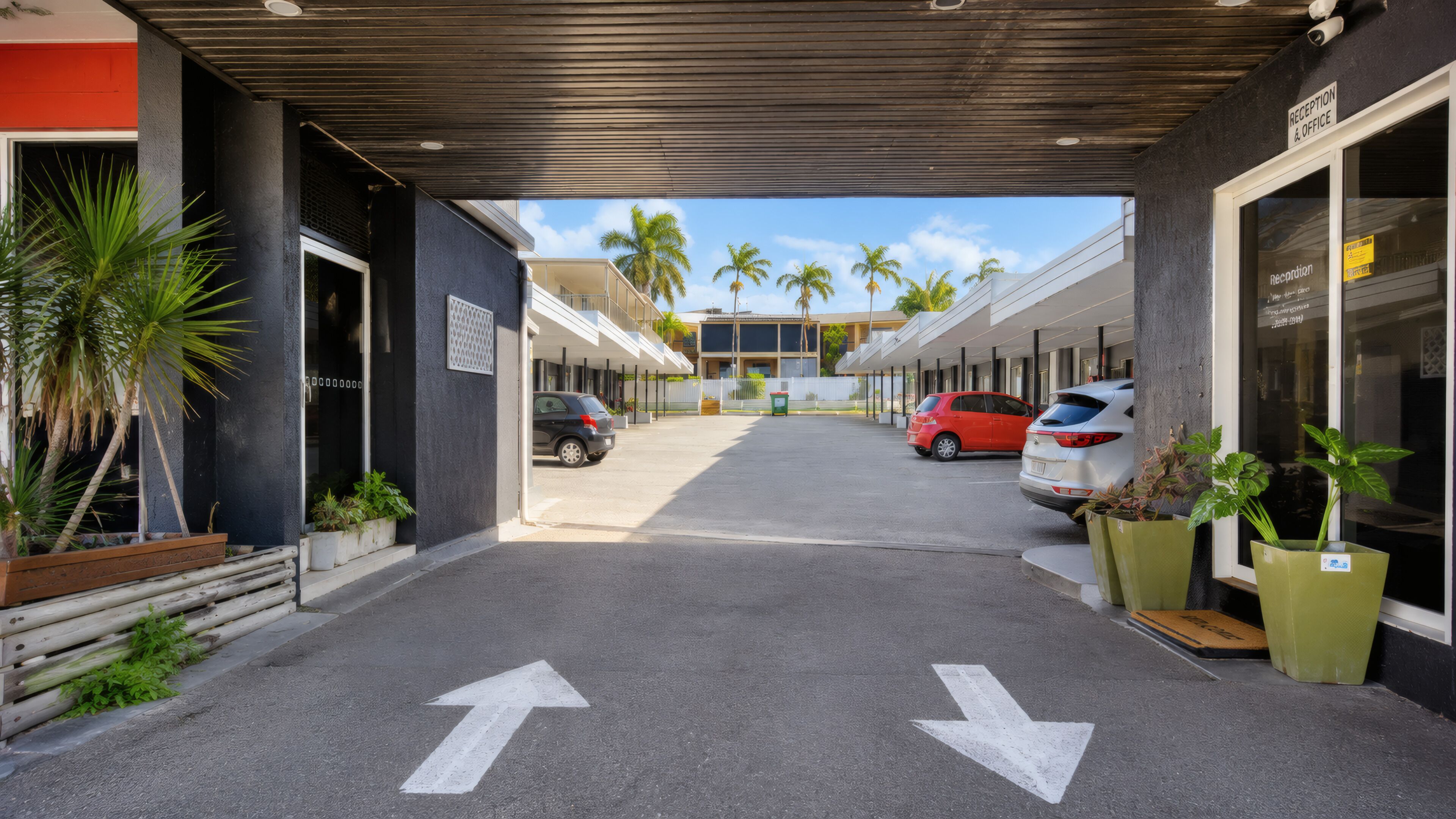 A covered walkway leading to a building with a reception office, palm trees, and parked cars.