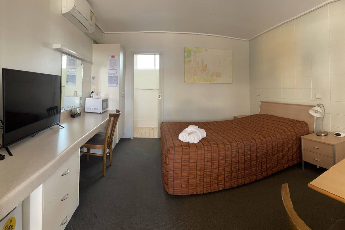 A compact hotel room with a bed, a desk with a television, a chair, a small table, and a nightstand.
