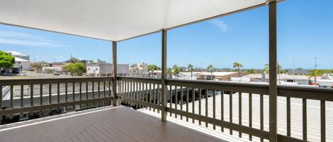 A covered outdoor area with a wooden deck, metal railing, and a view of buildings and palm trees.
