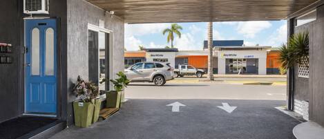Fachada de un edificio con puerta azul, plantas en macetas y un área de estacionamiento con un auto, y una tienda Ray White al fondo.