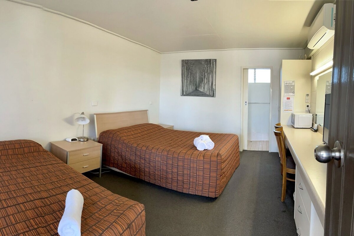 Habitación de hotel con dos camas, una mesita de noche con lámpara, microondas y puerta de acceso a un baño.