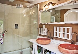 Une salle de bain dotée d’une douche à parois de verre, de deux vasques avec des cuves en céramique rondes, d’un grand miroir et d’un meuble-lavabo mural agrémenté d’une grille composée de petites étagères.