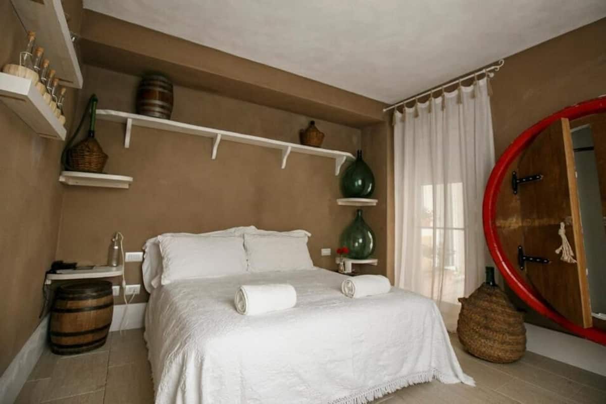 Un dormitorio con cama, espejo redondo, estantes con adornos y puerta de madera.