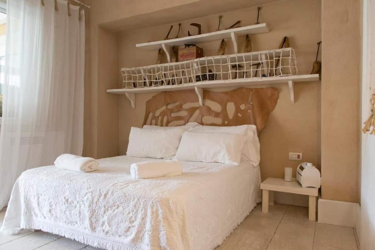 Una cama bien hecha con sábanas blancas, una mesita de noche con lámpara, y repisas sobre la cama con objetos decorativos.