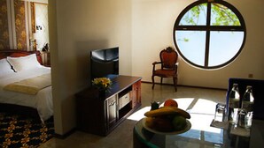 A hotel room with a bed, a television, a chair, a round window, and a table with fruits and bottles. - Cron Palace Tbilisi Hotel (Tbilisi)