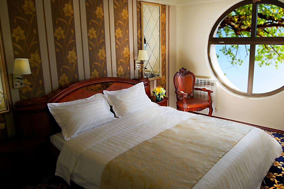 A neatly made bed with white linens, a wooden headboard, and a floral-patterned wall. There is a round window with a view of trees and a chair with a vase of flowers.