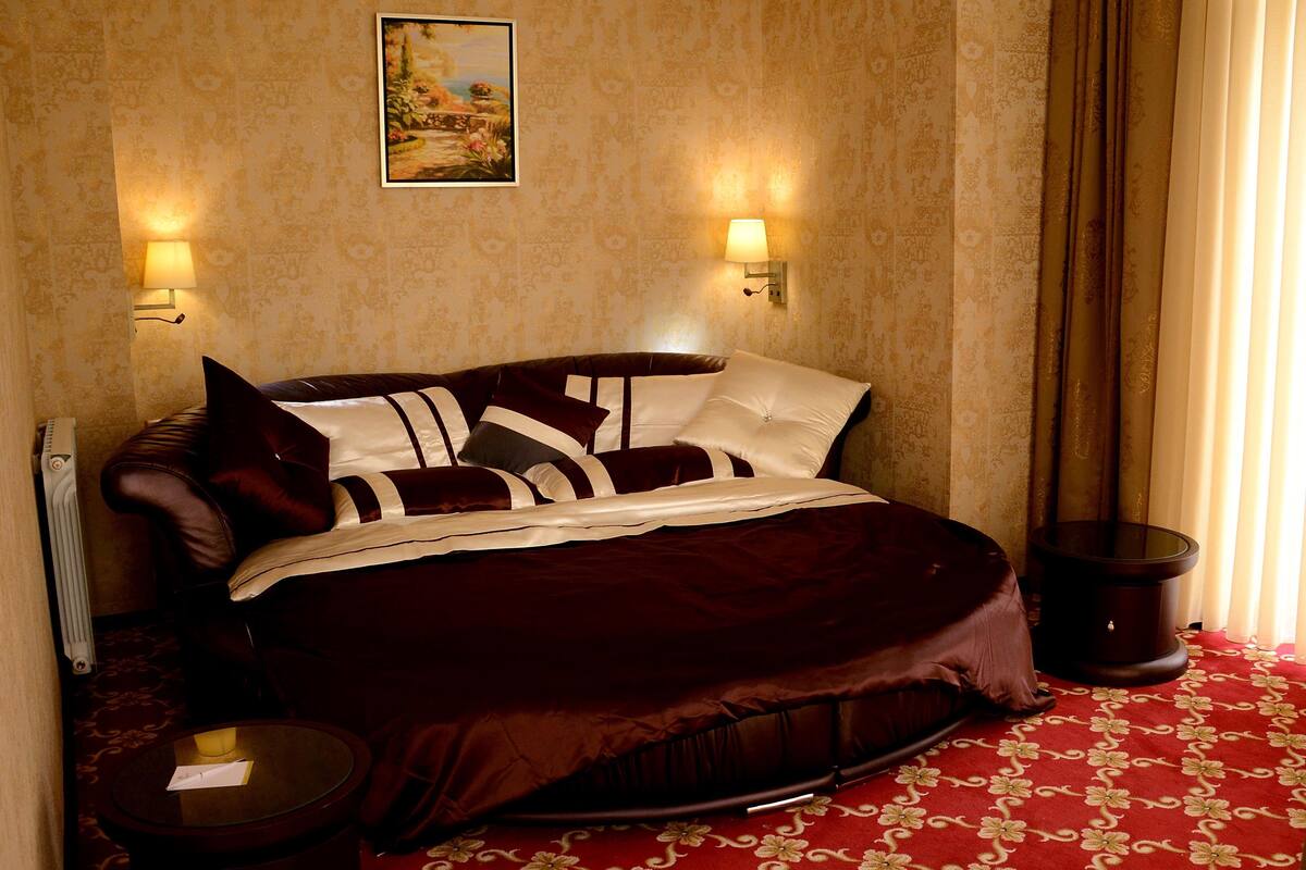 A room with a large bed, two wall-mounted lamps, a radiator, and a carpeted floor.