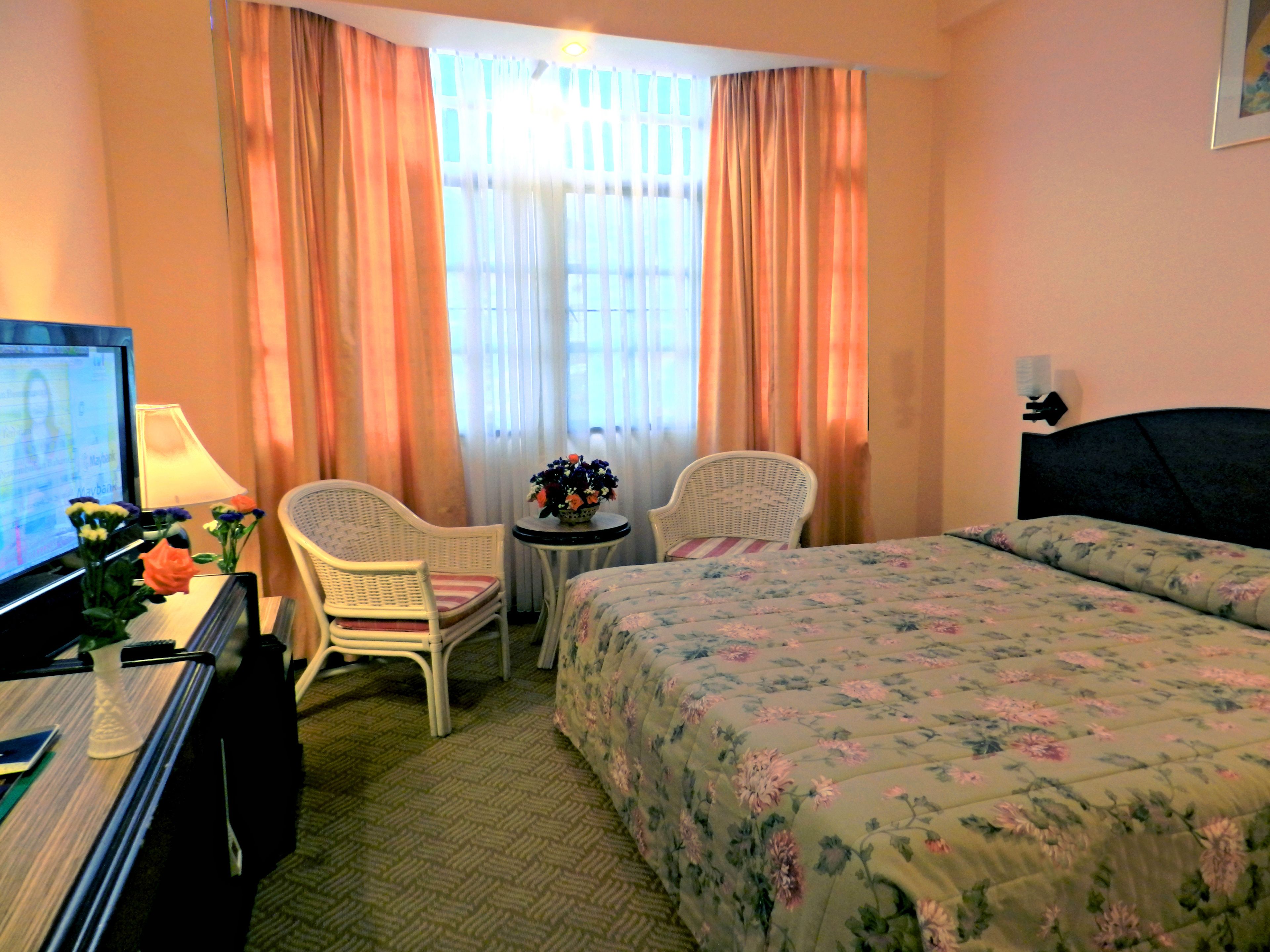 A hotel room with a bed, two chairs, a small table with a vase of flowers, a desk with a computer, and a window with curtains.