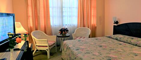 A hotel room with a bed, two chairs, a small table with a vase of flowers, a desk with a computer, and a window with curtains.