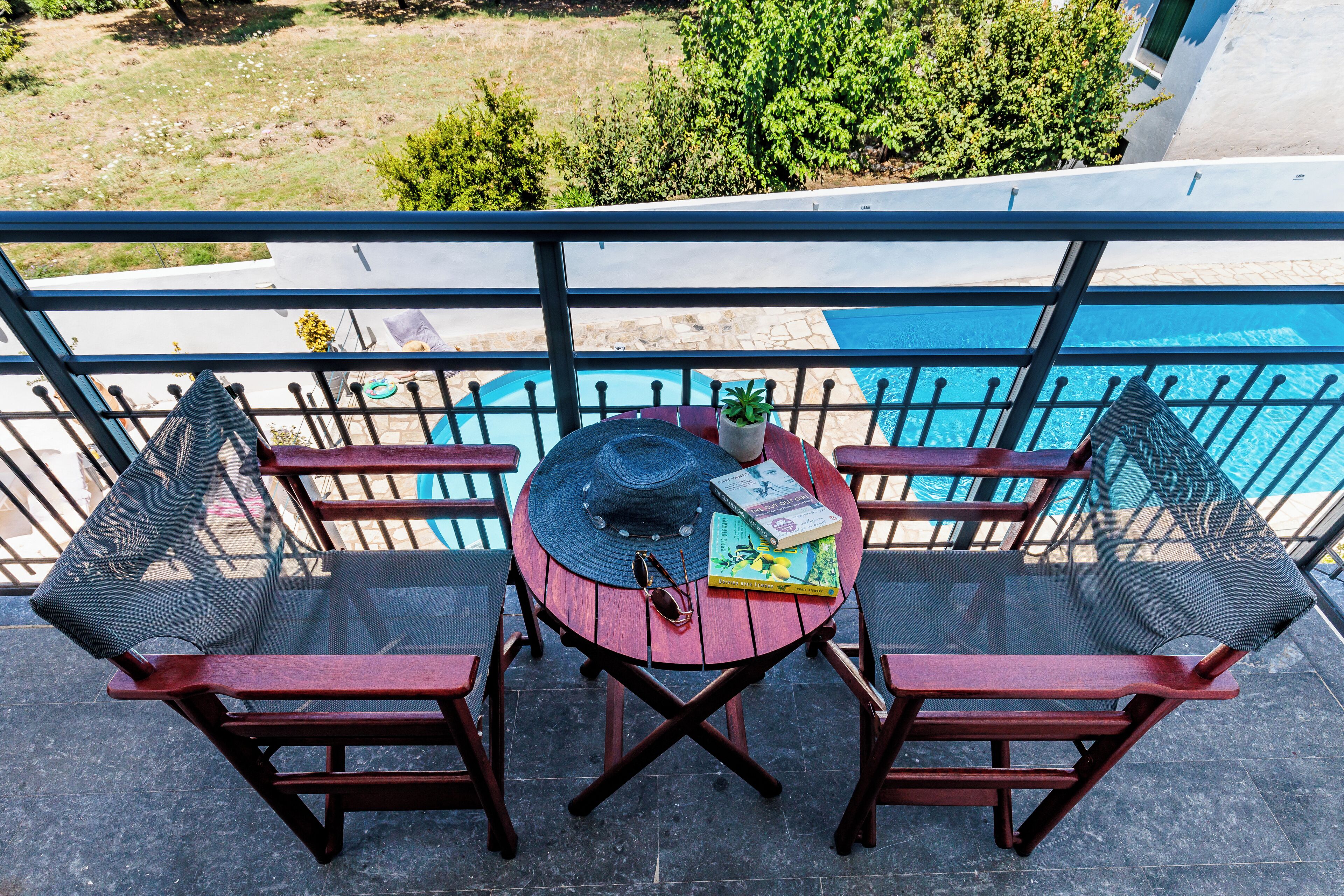 A round table with a hat, sunglasses, and books on a balcony with a pool view.