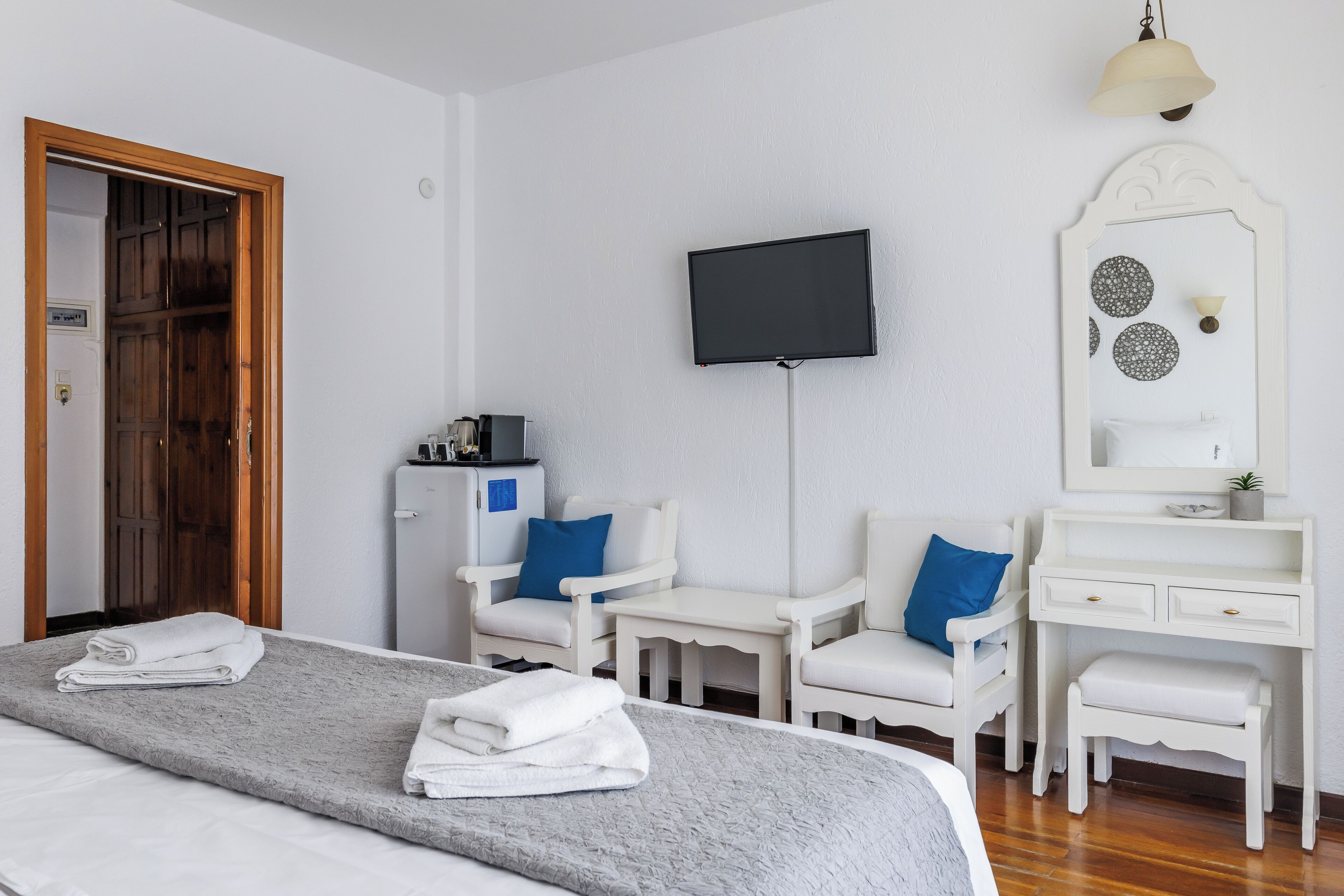 A hotel room with a bed, two white chairs, a desk, a television, and a mirror.