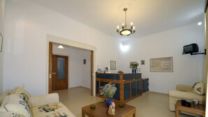 A living room with a sofa, a coffee table with a vase and a small statue, a wooden cabinet, and a television mounted on the wall. - Corali Hotel (Milos)