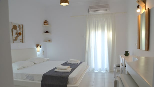 A bedroom with a bed, bedside tables, a mirror, and a window with curtains. - Corali Hotel (Milos)