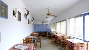 A dining area with checkered tablecloths, wooden chairs, and a ceiling fan. - Corali Hotel (Milos)