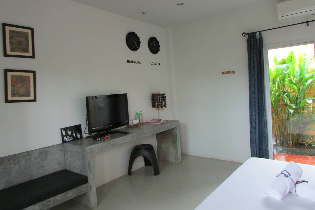 A modern hotel room with a flat-screen TV, a small desk with a computer, a bed with a rolled towel, and a view of greenery through the window.