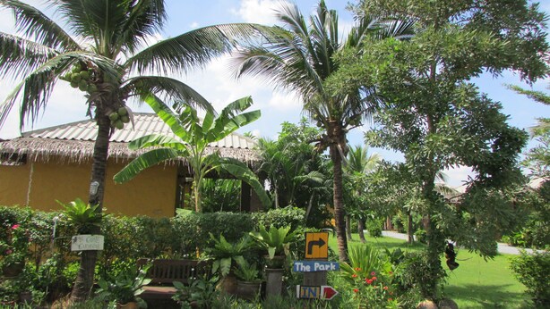 A tropical garden with palm trees, a thatched-roof building, and directional signs. - The Park Drive-Thru Check-In Resort (Thanyaburi)