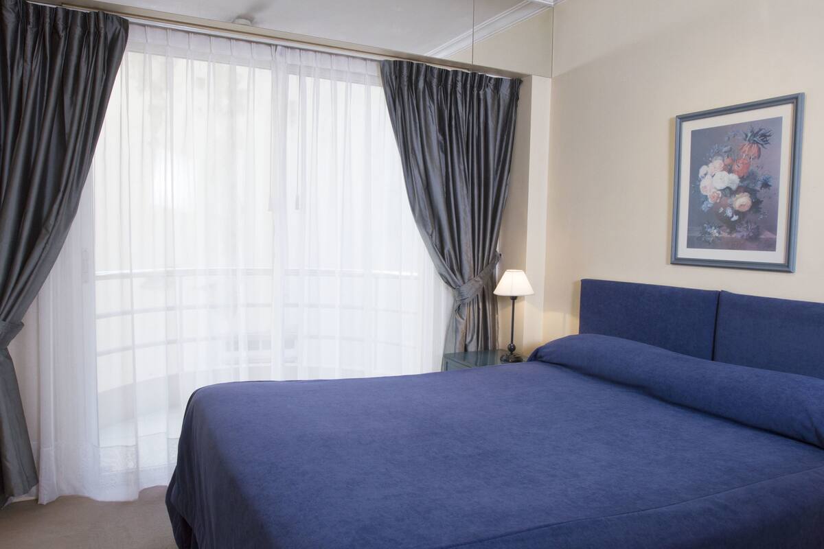 Une chambre à coucher avec un grand lit, une table de chevet avec une lampe et une fenêtre avec des rideaux.