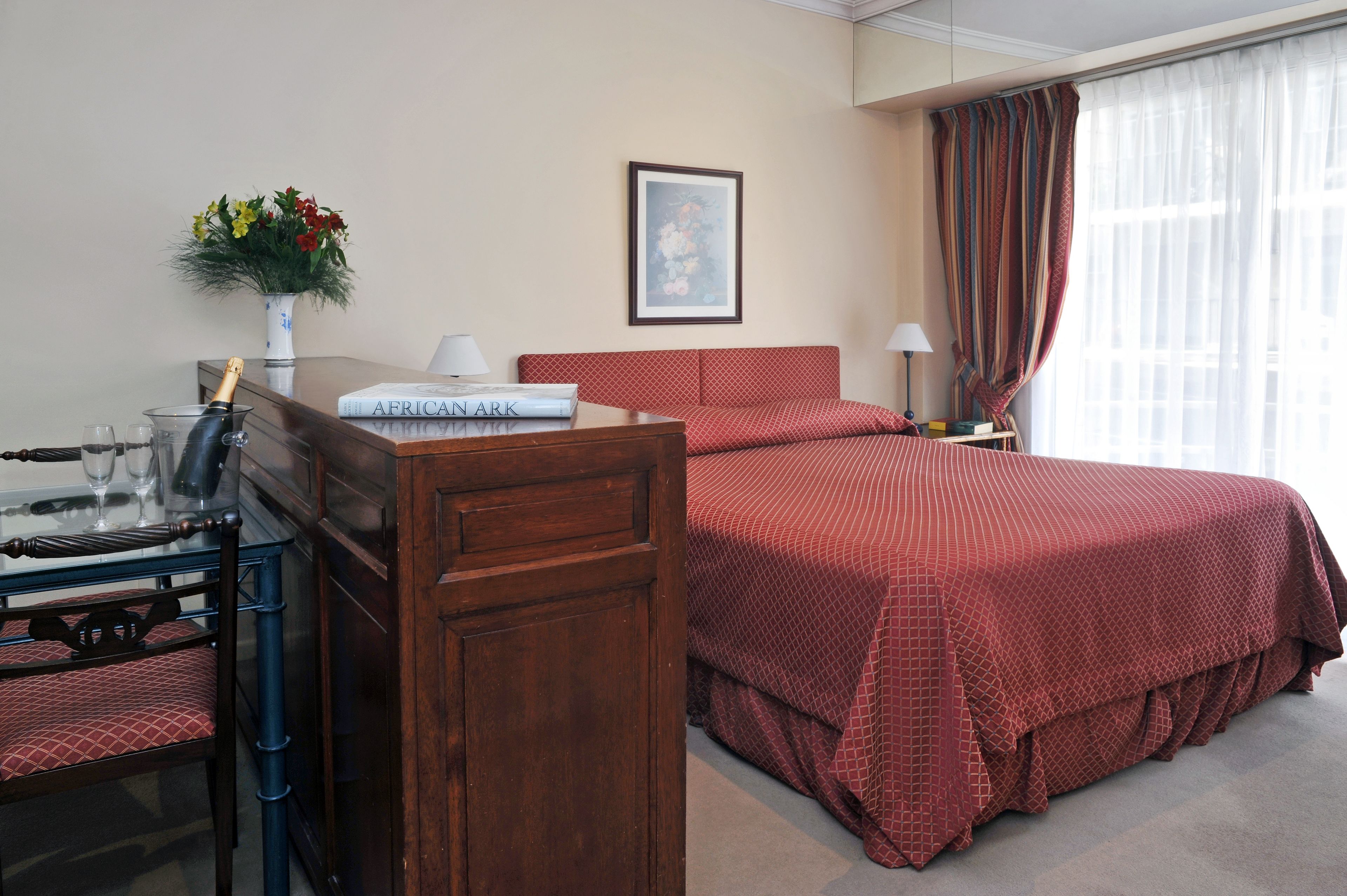 A hotel room with a wooden desk, a bed with red bedding, a vase with flowers, a framed picture on the wall, and a window with curtains.