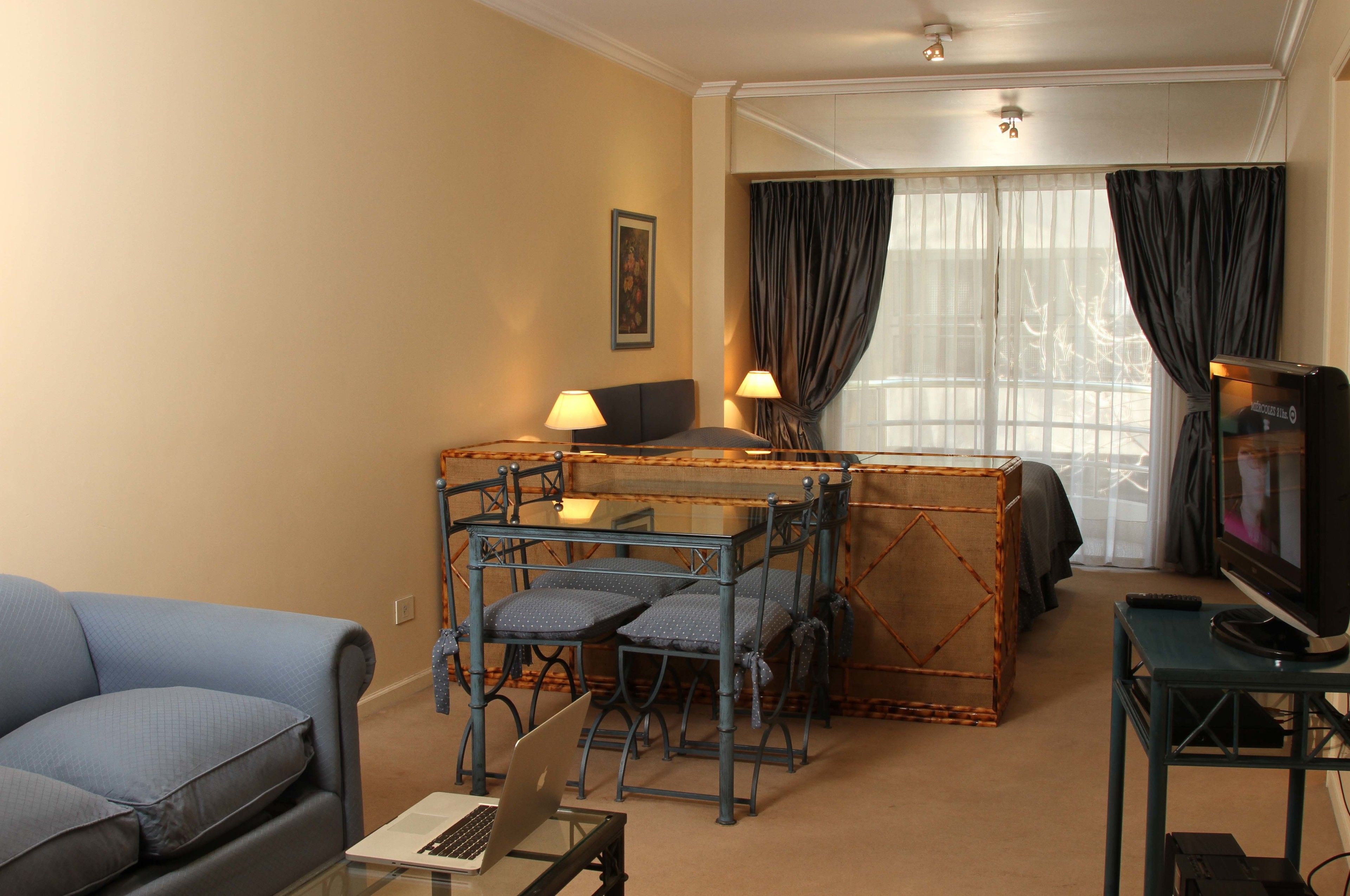 A living room with a sofa, a small table with a laptop, a TV, and a dining area with chairs and a table.