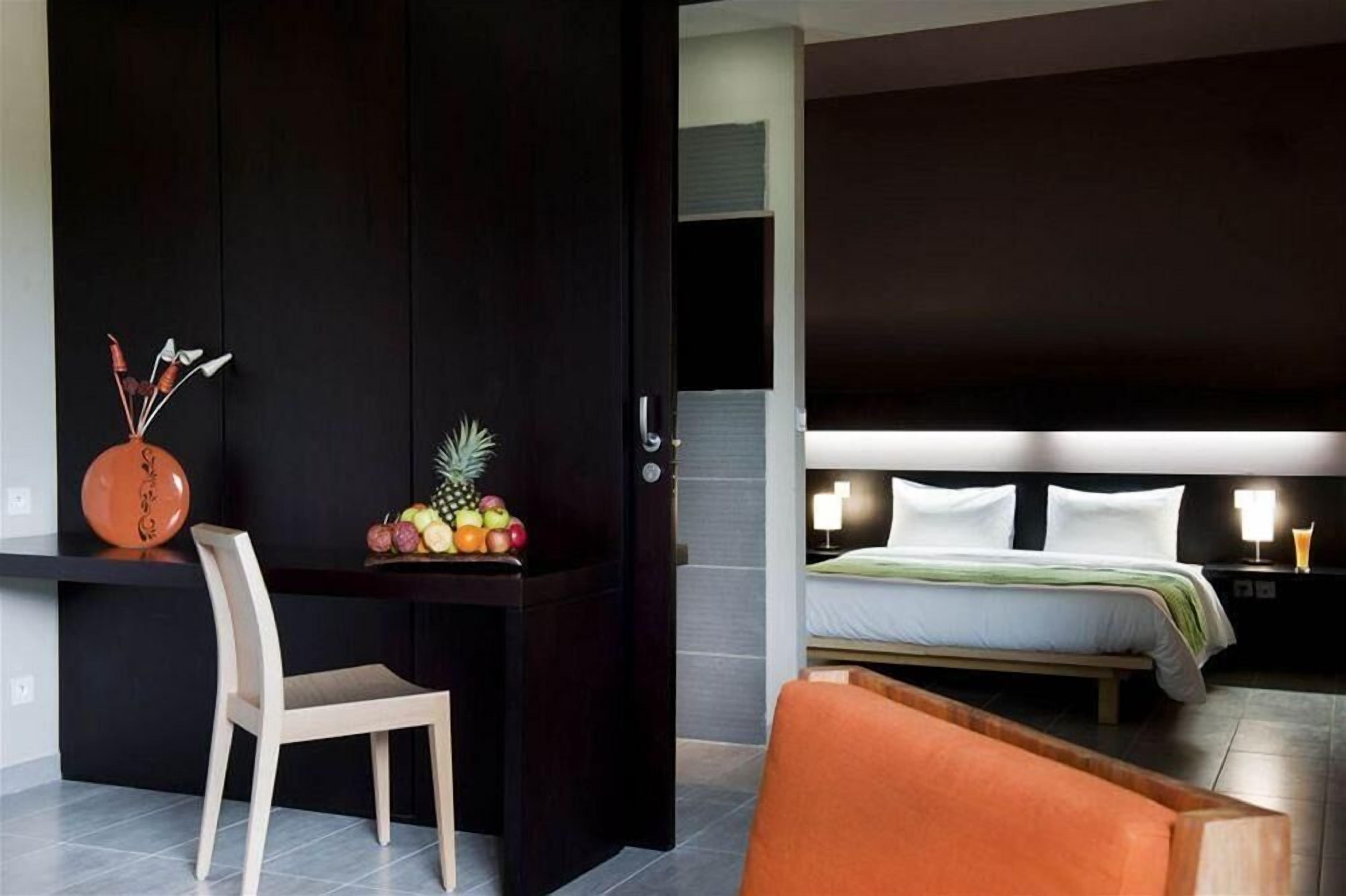 Una habitación de hotel con cama, mesita de noche, silla y escritorio con fruta.