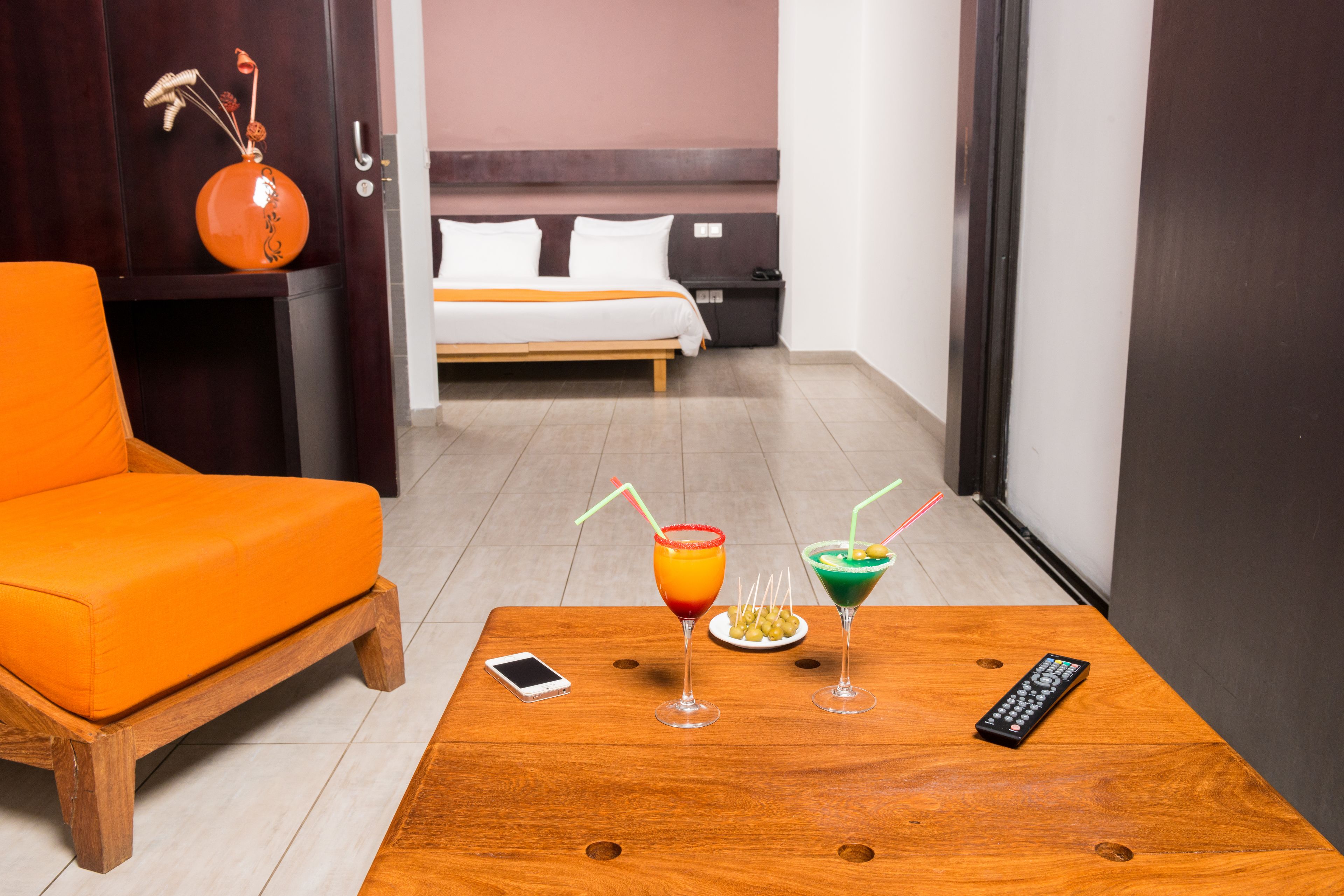 Une chambre d’hôtel avec une table en bois, un canapé orange et un lit avec une literie blanche. Il y a deux boissons sur la table et une télécommande.