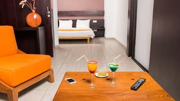 Une chambre d’hôtel avec une table en bois, un canapé orange et un lit avec une literie blanche. Il y a deux boissons sur la table et une télécommande.