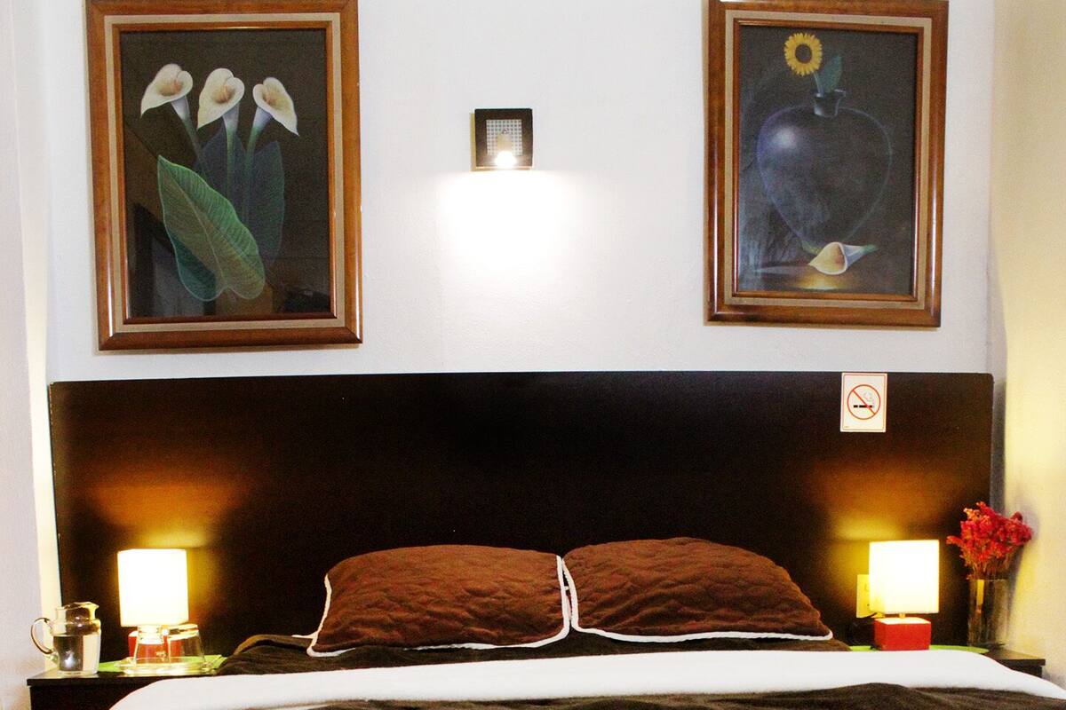 Una cama bien hecha con cabecera marrón, dos lámparas de noche y cuadros en la pared.