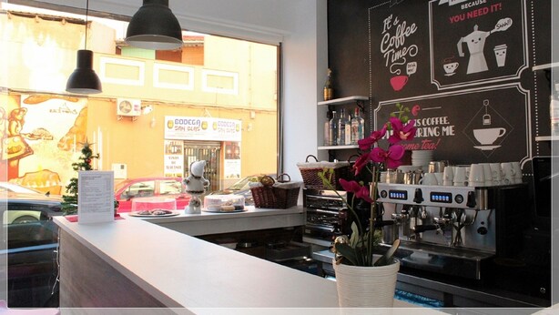 A coffee shop interior with a counter, espresso machine, hanging lights, and a wall with coffee-related graphics. - Chameleon Hostel (Alicante)