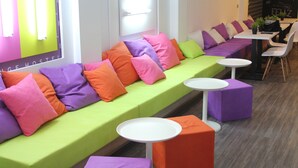 A modern lounge area with a long green sofa adorned with colorful cushions, white round tables, and orange and purple cube-shaped stools. - Chameleon Hostel (Alicante)
