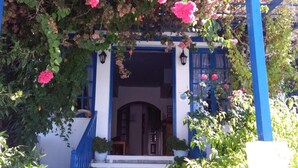 Fachada azul y blanca de un edificio, un conjunto de escalones de piedra que conducen a una puerta, y una pérgola cubierta de flores rosadas.