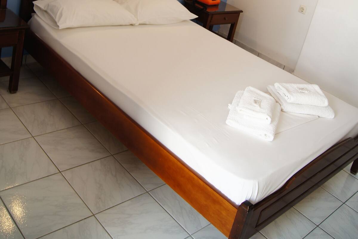 A neatly made bed with white linens and two pillows, a wooden headboard, two bedside tables with lamps, and a small orange bag on one table.