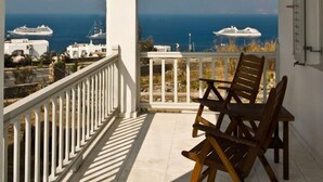 Ein Balkon mit Blick auf das Meer und Schiffe, mit weißer Metallreling und Holzstühlen.