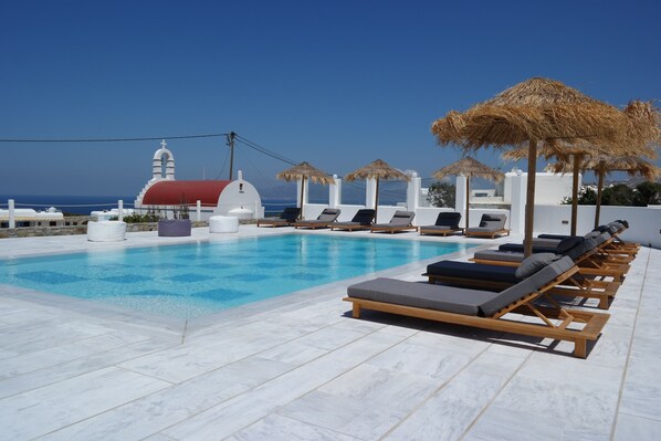 Una piscina en la azotea con sillones y sombrillas, un edificio blanco con techo rojo y un cielo azul despejado.