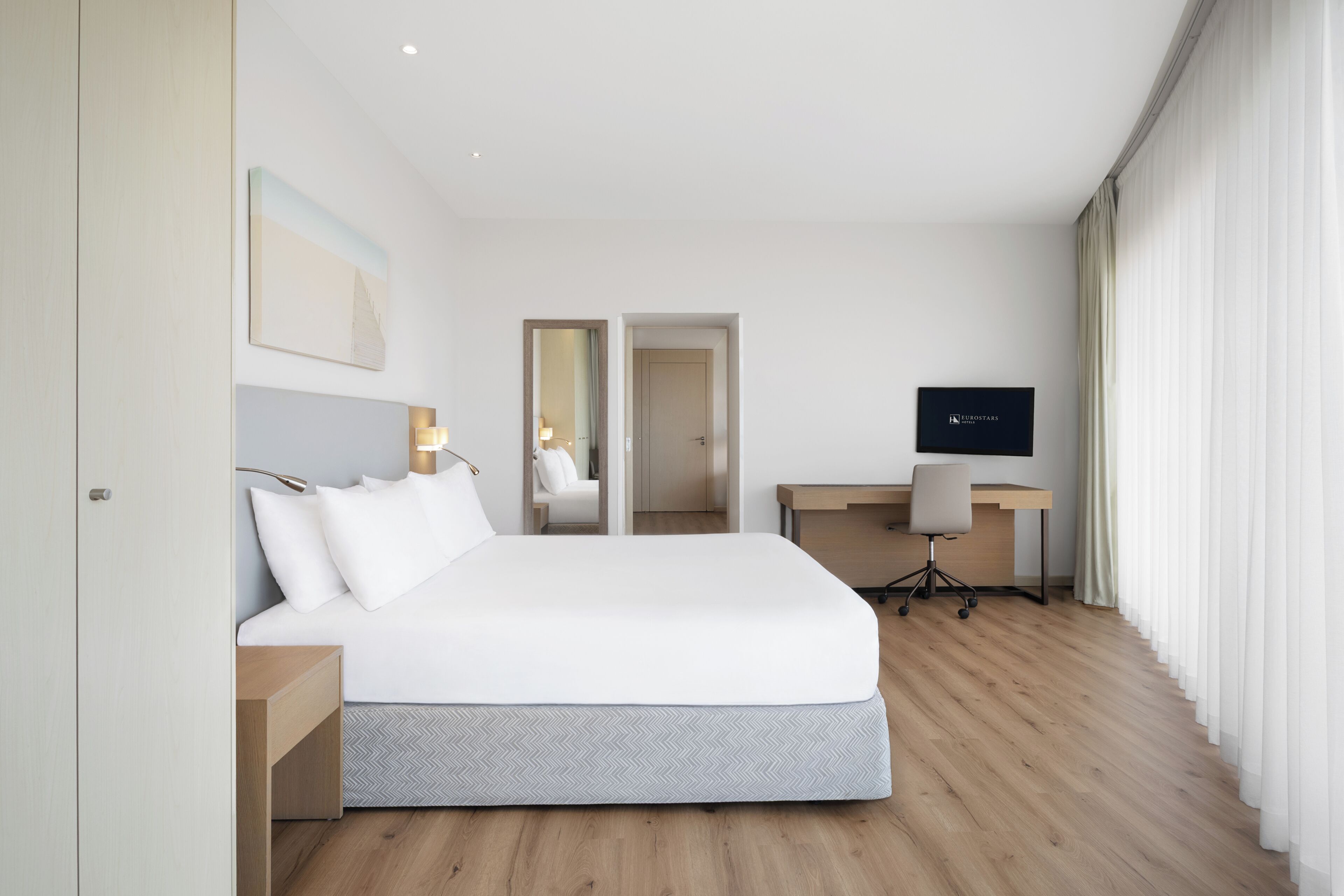 A modern hotel room with a large bed, a desk, a chair, and a television.