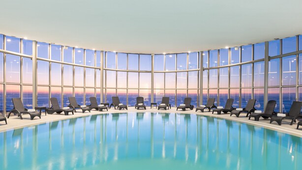A large indoor swimming pool with lounge chairs and large windows offering a view of the sunset over the ocean.