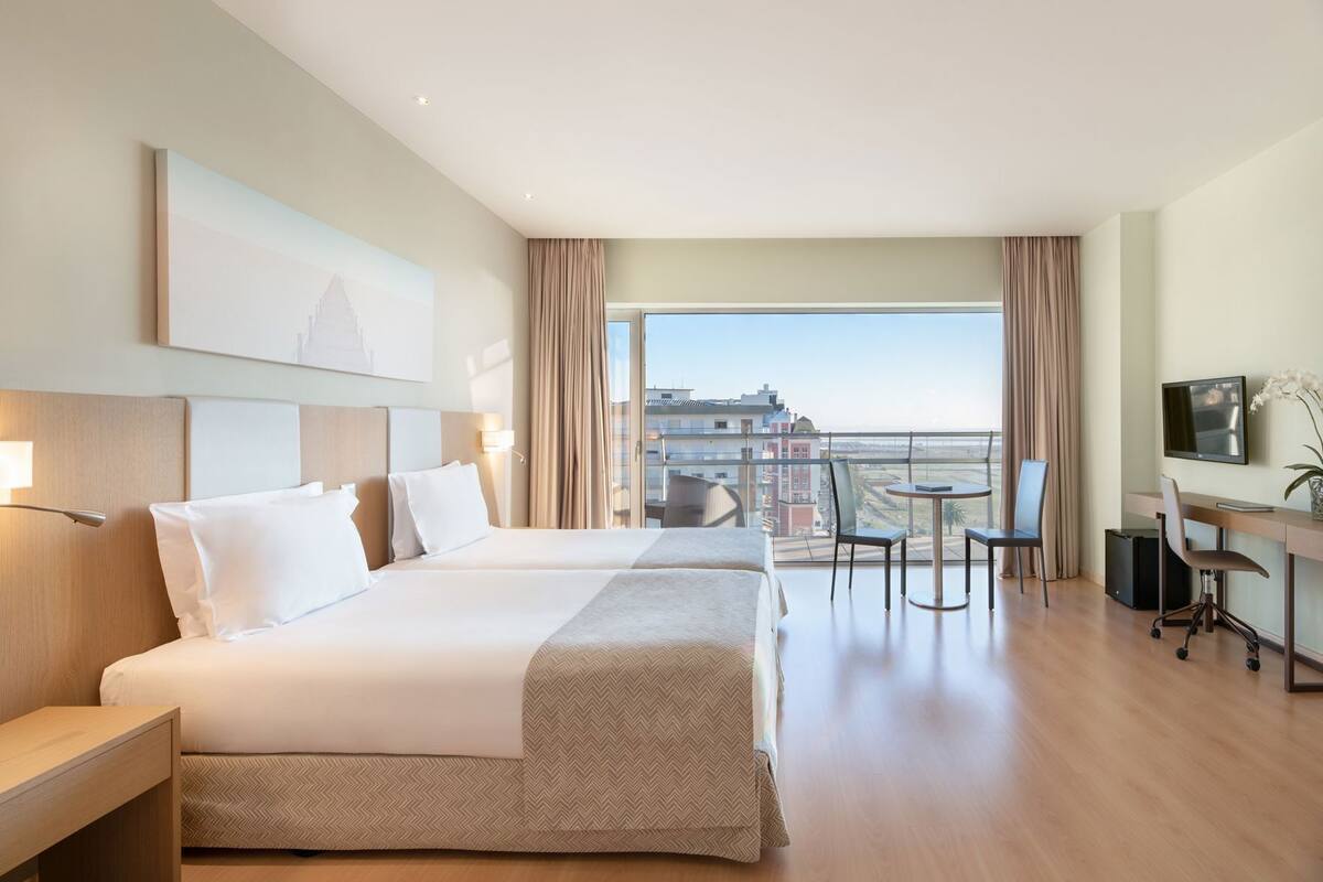 A hotel room with two beds, a desk with a chair, a TV, and a balcony with a view of the city.
