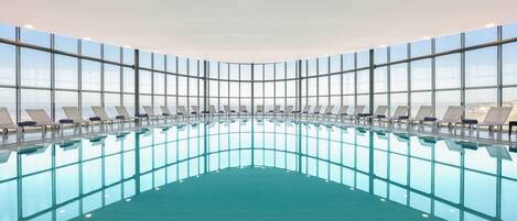 Une grande piscine intérieure aux parois en verre transparent, entourée de chaises longues.