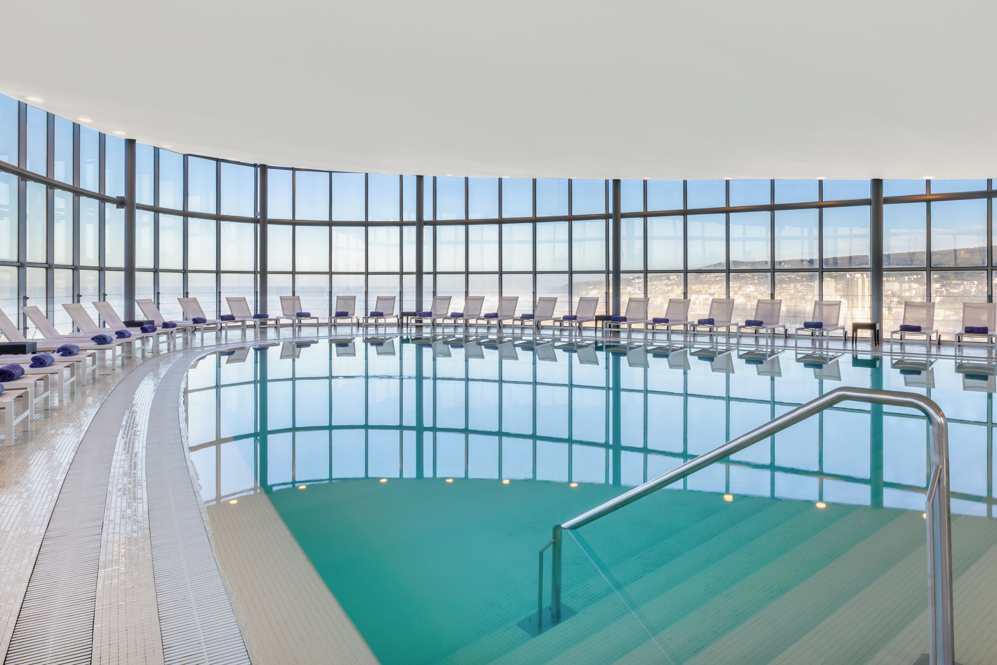 A large indoor swimming pool with clear blue water, surrounded by lounge chairs and a high ceiling with large windows.