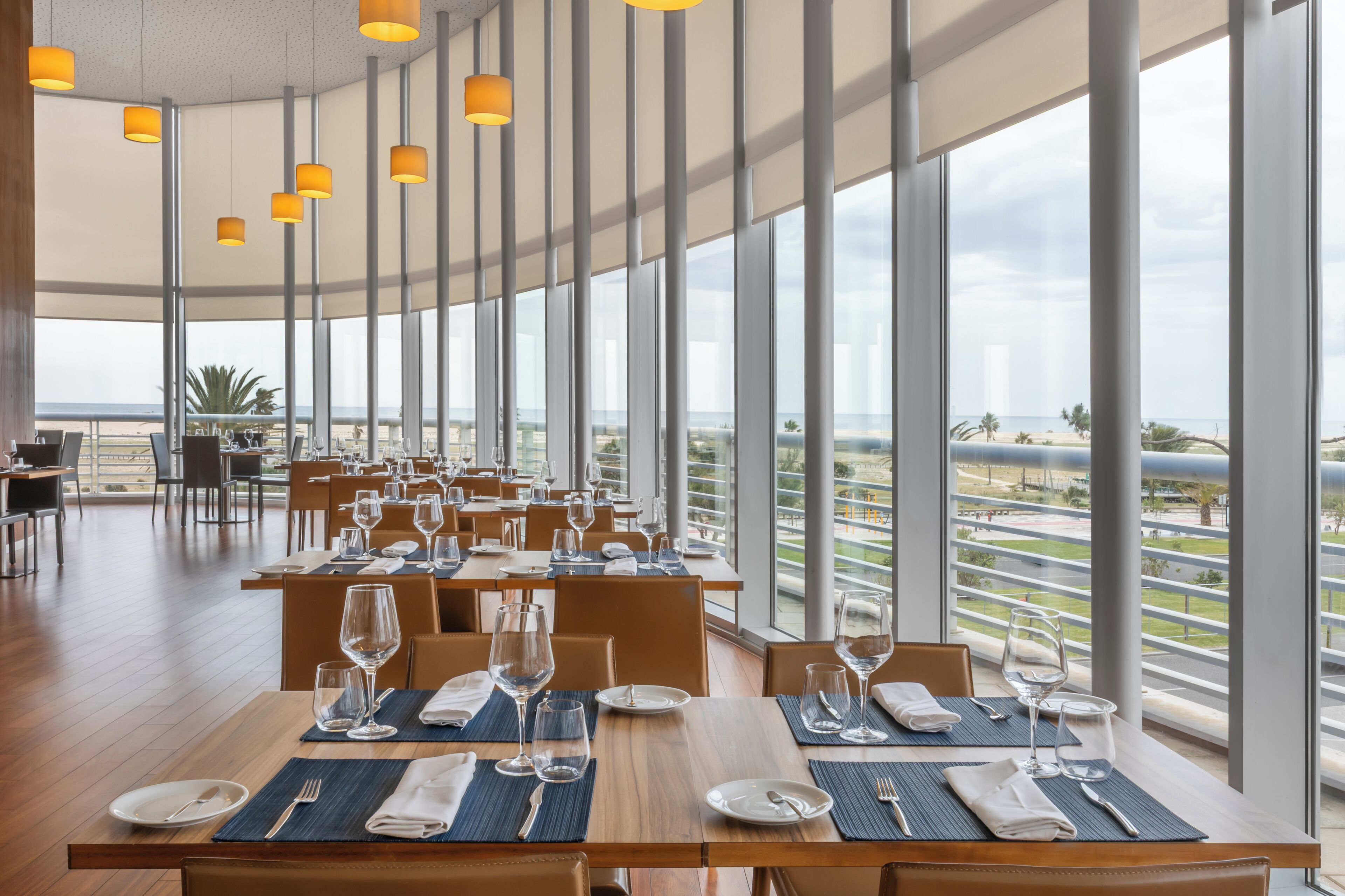 A restaurant with tables set for dining, large windows offering a view of the beach, and hanging pendant lights.