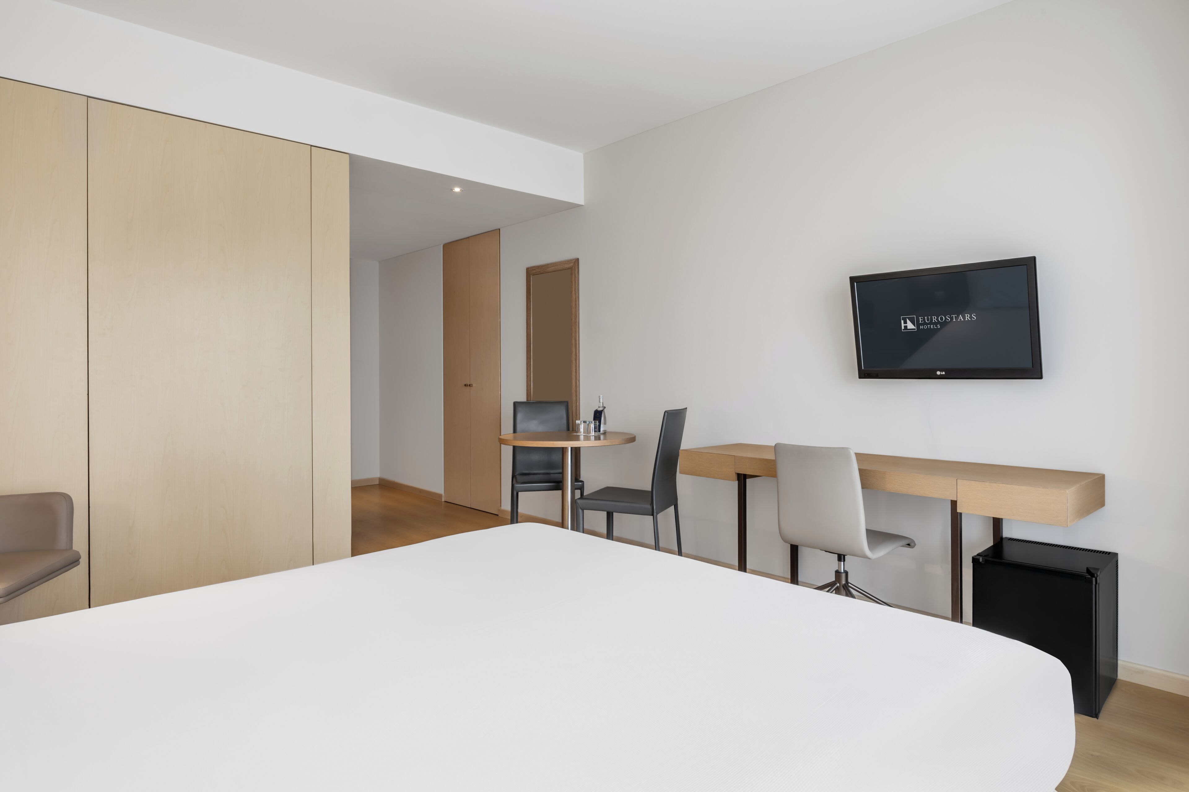 A modern hotel room with a bed, a desk, two chairs, a television, and a trash bin.