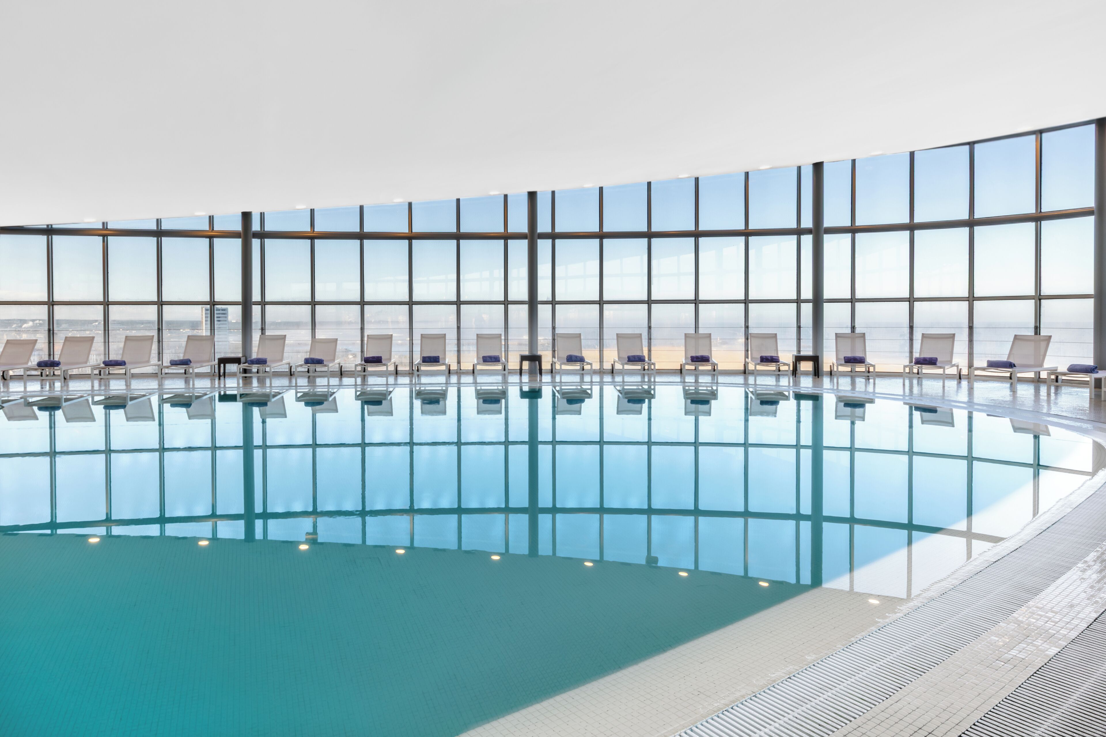 An indoor swimming pool with clear glass walls, lounge chairs, and a tiled floor.