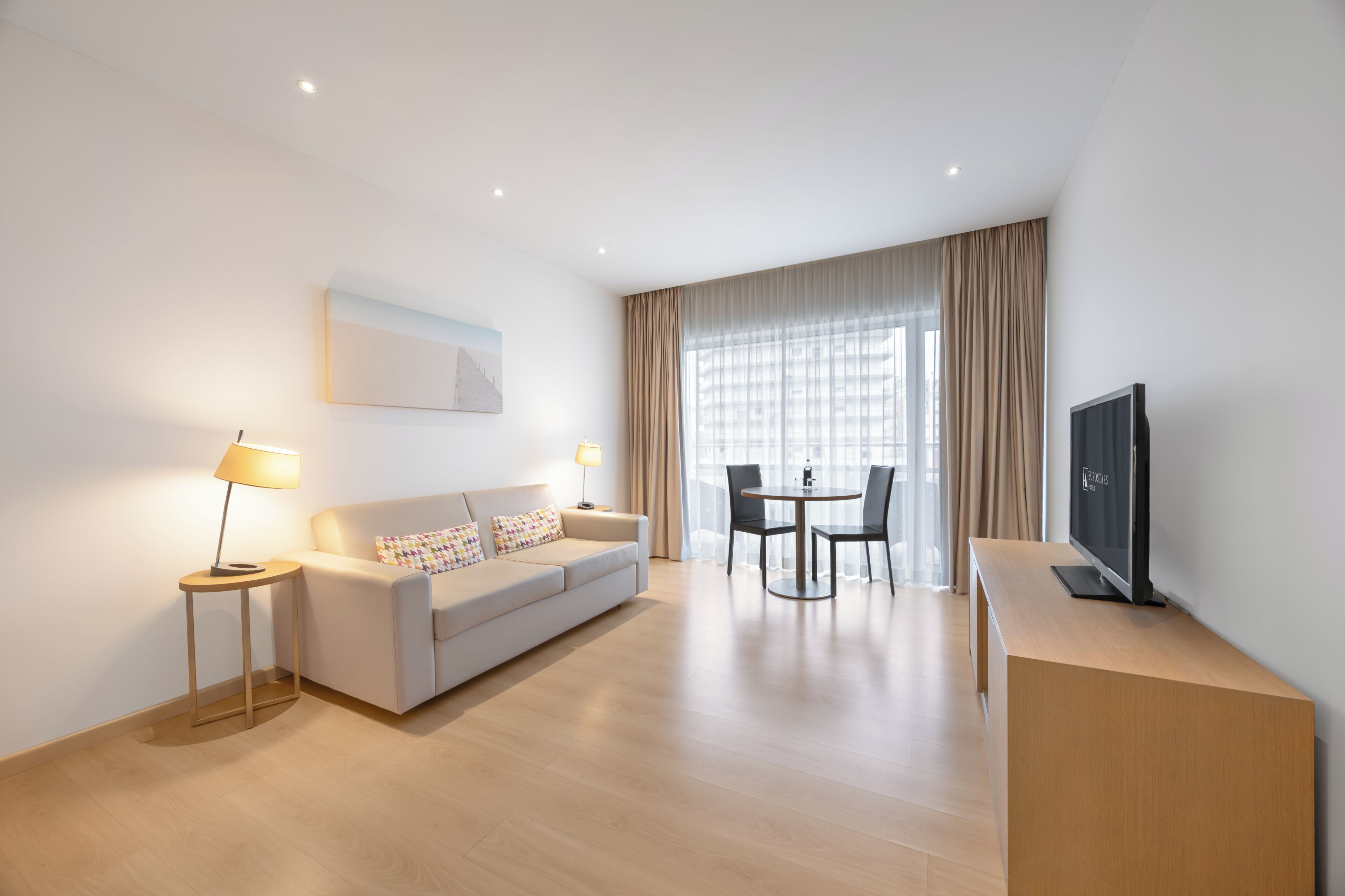 Ein modernes Hotelzimmer mit einer Couch, einem Esstisch mit Stühlen, einem Fernseher und einem fenster mit Vorhängen.