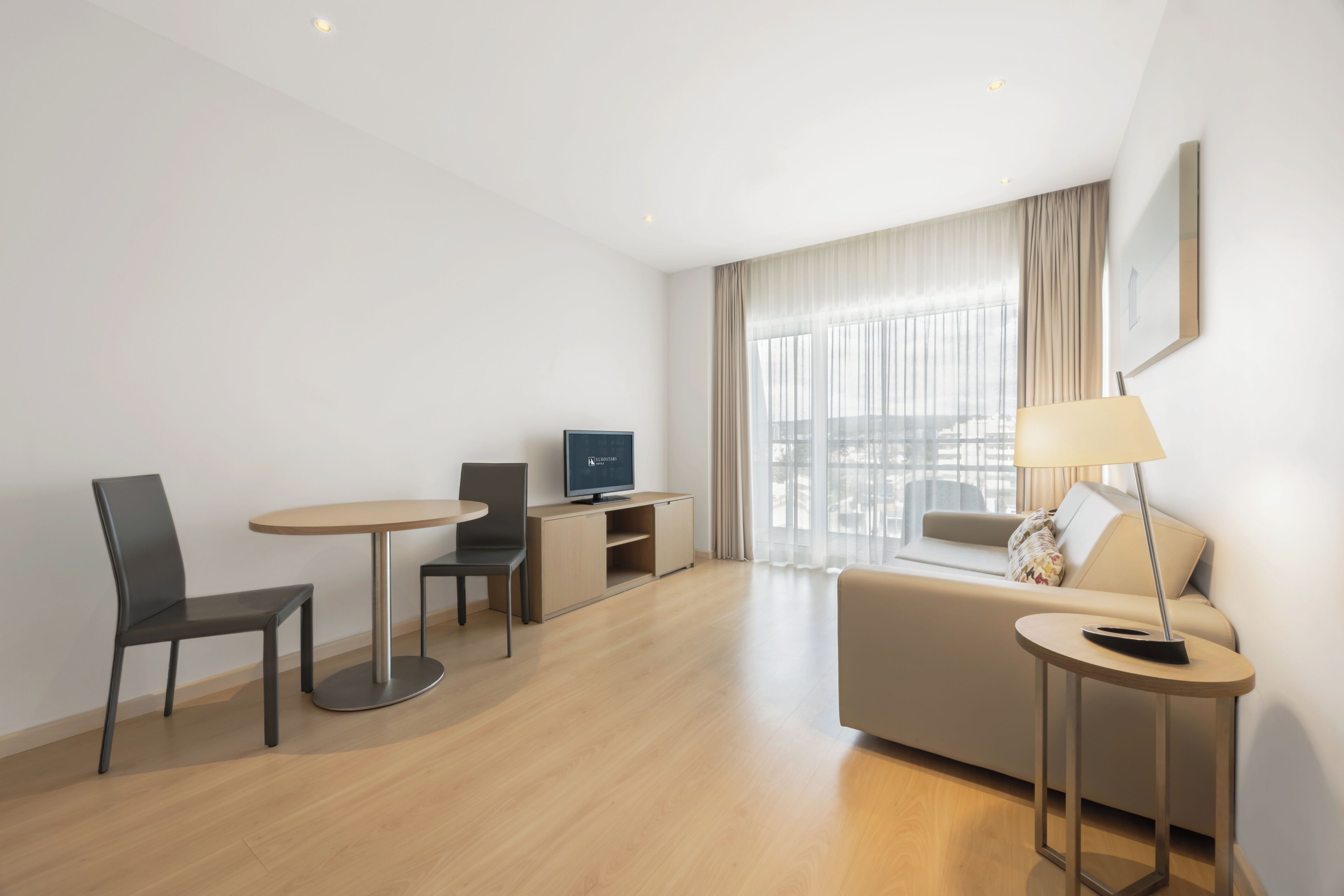 Quarto de hotel moderno com sofá, uma mesinha redonda, televisão e luminária.