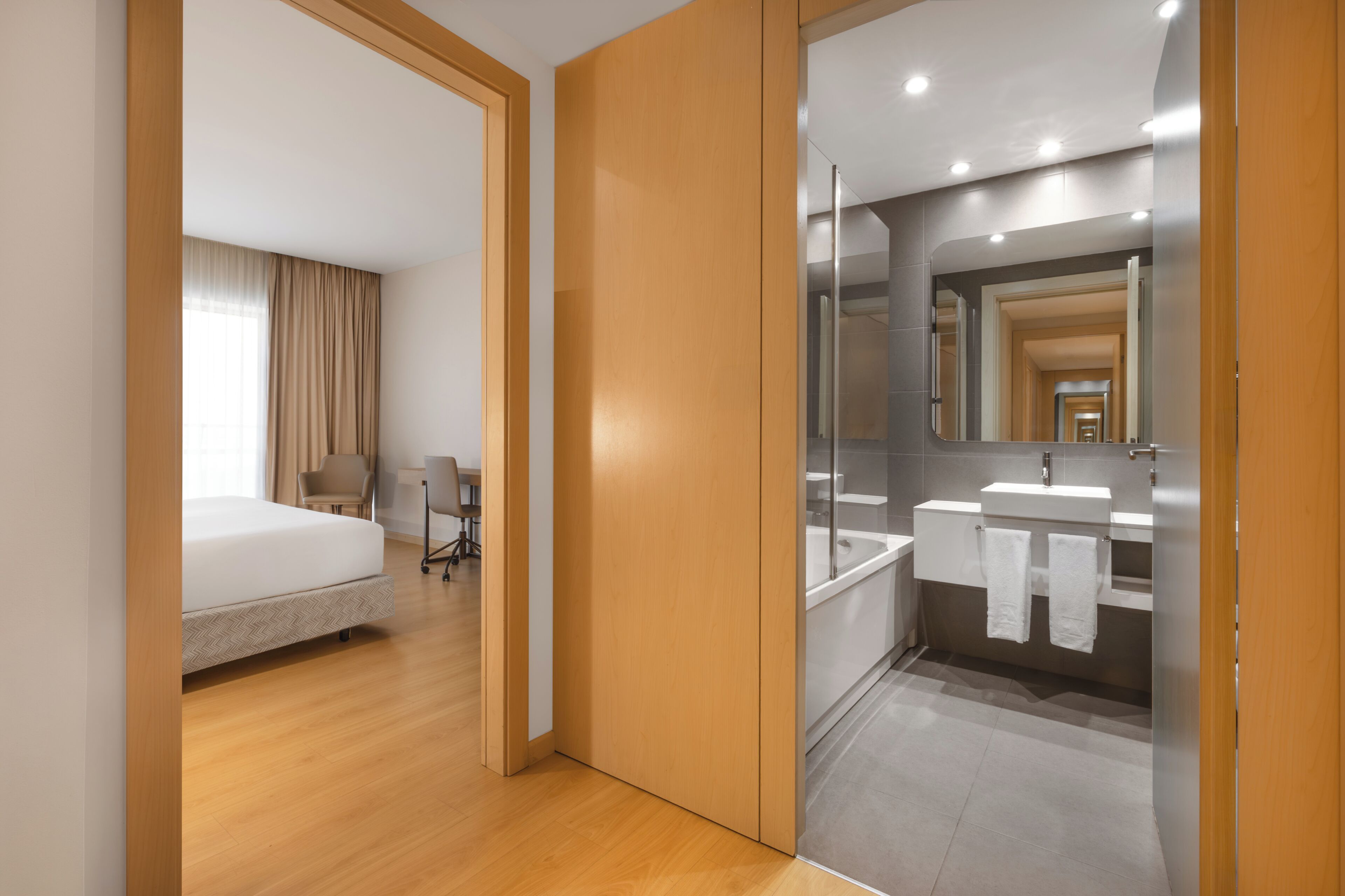 Una habitación de hotel moderna con cama, escritorio y baño con espejo, lavamanos y toallero.