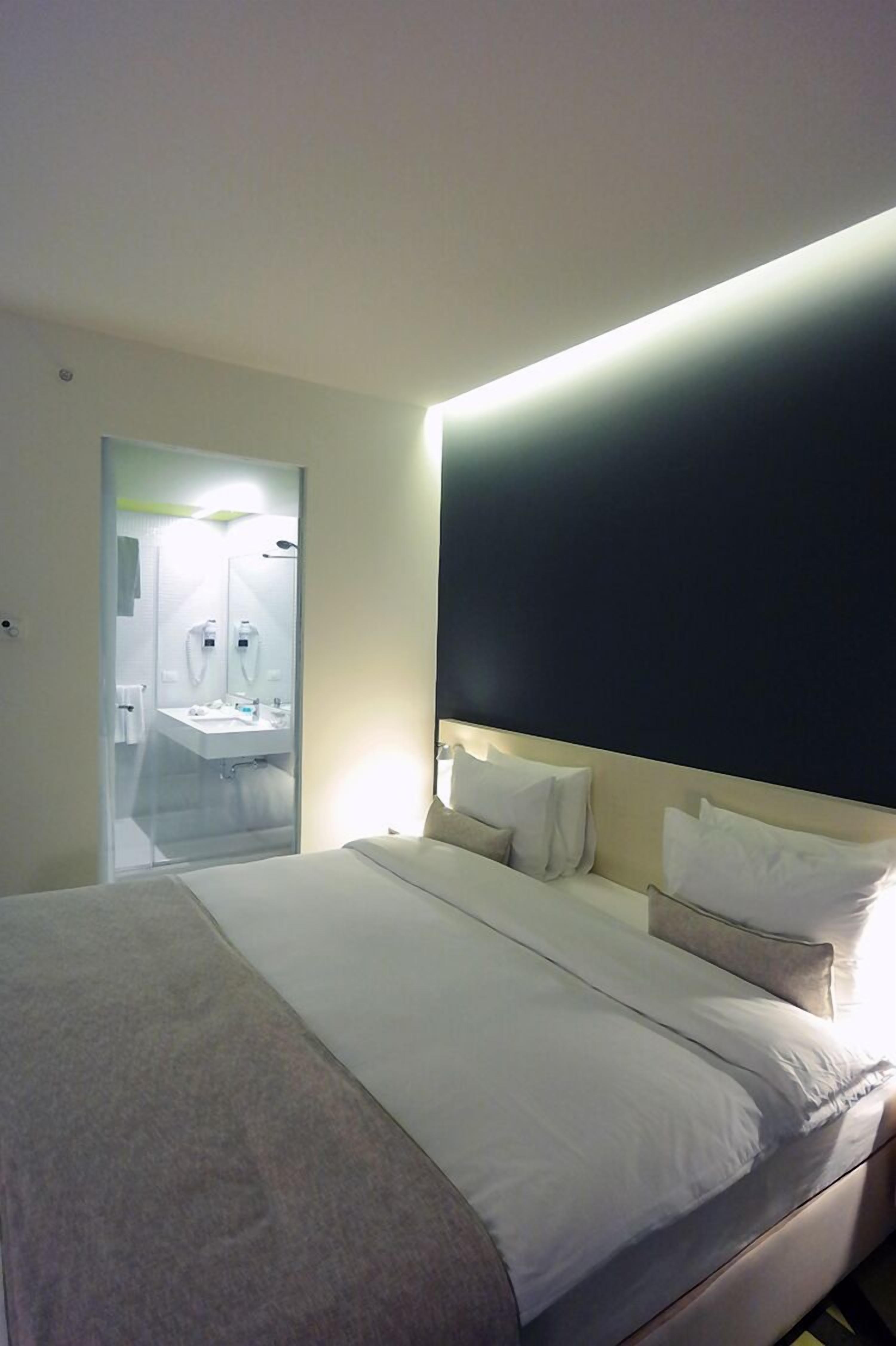 A hotel room with a bed, a bedside table, a large mirror, and a bathroom visible through a glass door.