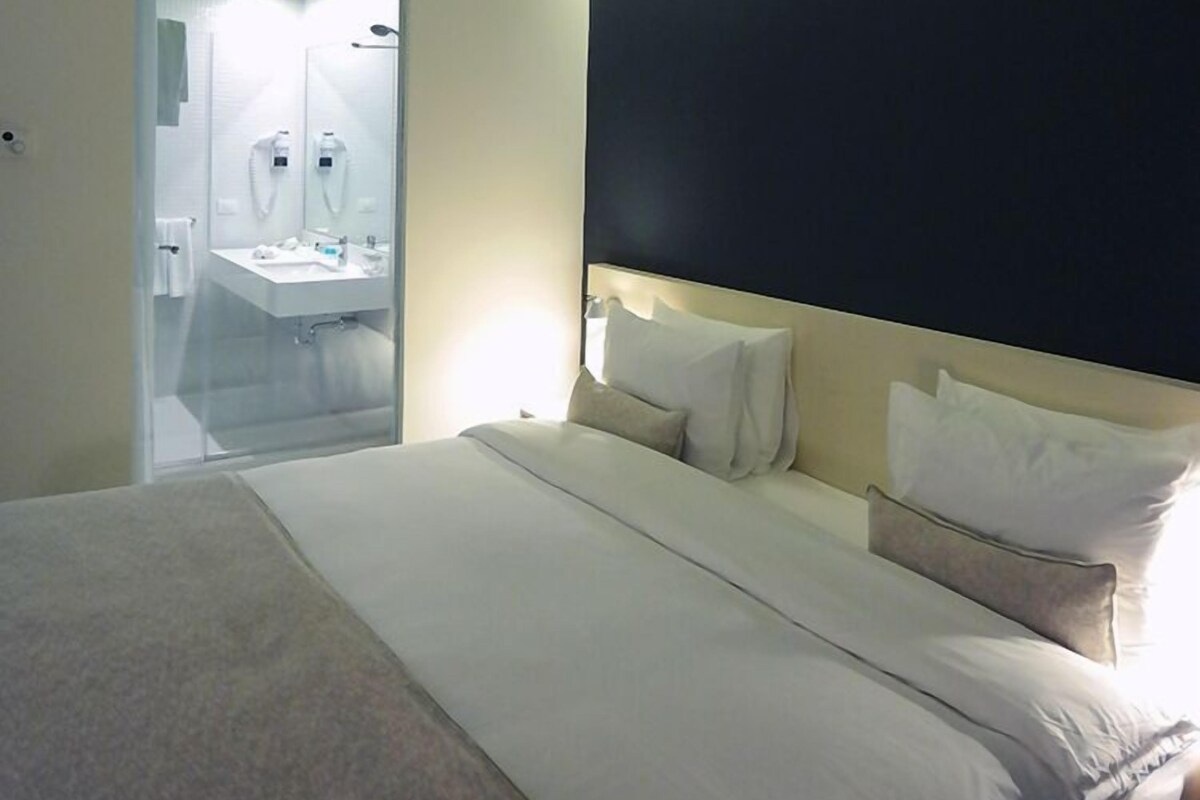 Habitación de hotel con cama, mesita de noche, un espejo grande y un baño visible a través de una puerta de cristal.