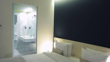 A hotel room with a bed, a bedside table, a large mirror, and a bathroom visible through a glass door.