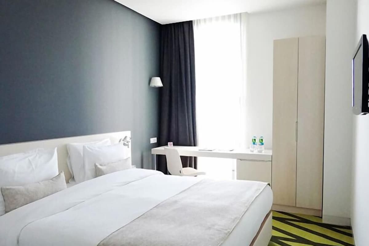 A hotel room with a bed, a desk, a chair, a wardrobe, and a security camera.