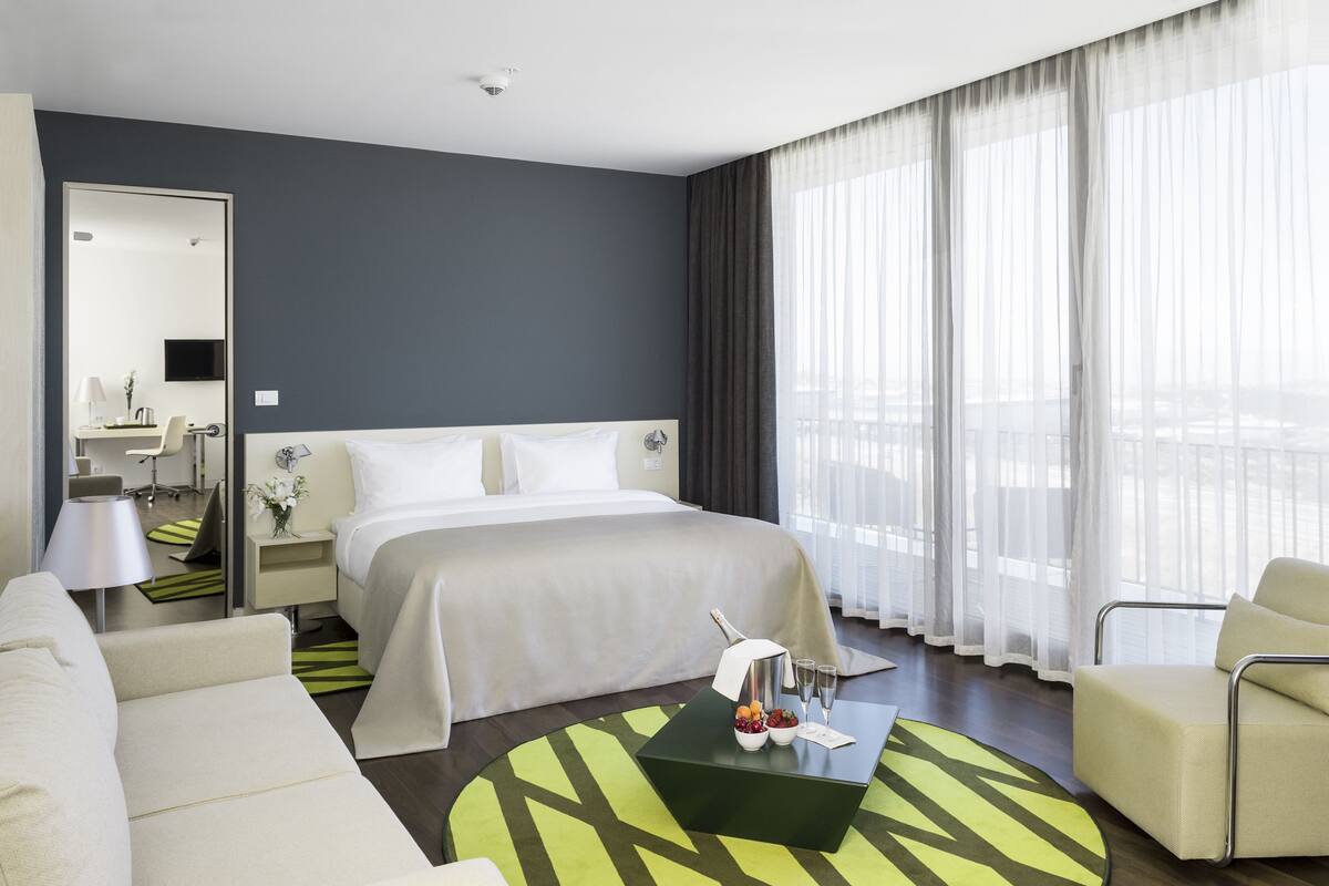 A hotel room with a large bed, a sitting area with a sofa and armchair, a small table with a bottle and glasses, and a large window with sheer curtains.
