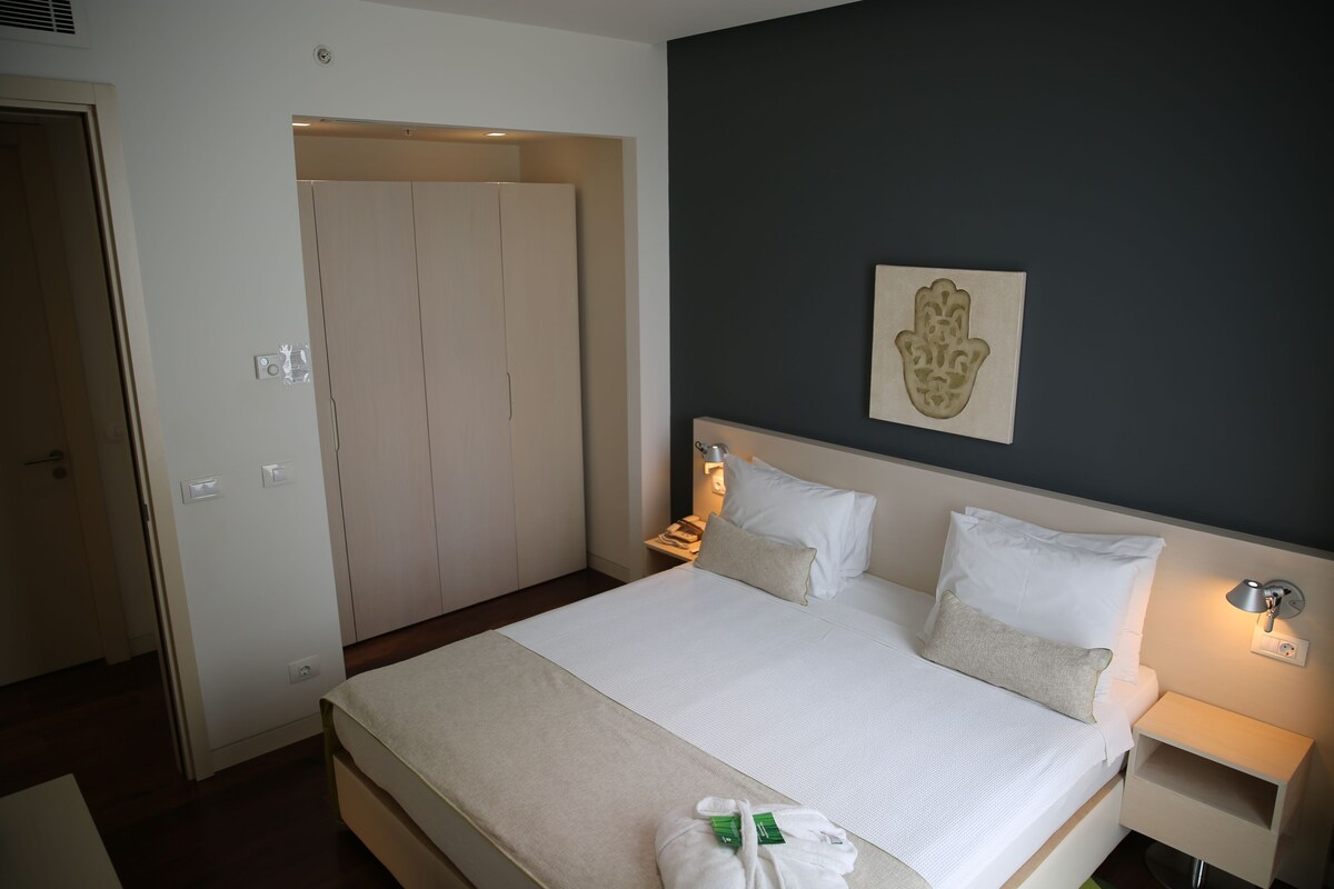 A hotel room with a bed, bedside lamps, a nightstand, a wall-mounted artwork, and a door leading to another room.